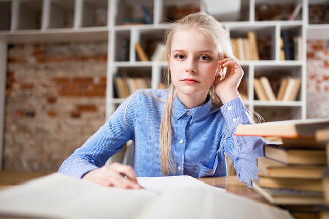 Mladé dievča s blond vlasmi sedí pri stole s kopou kníh.jpg